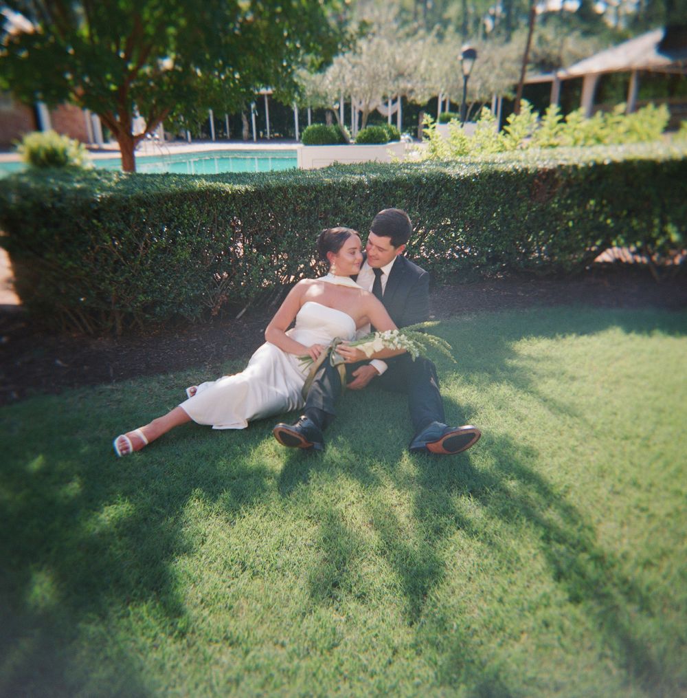A couple dips back into an embrace while posing on a sunny lawn with manicured hedges and trees in the background.