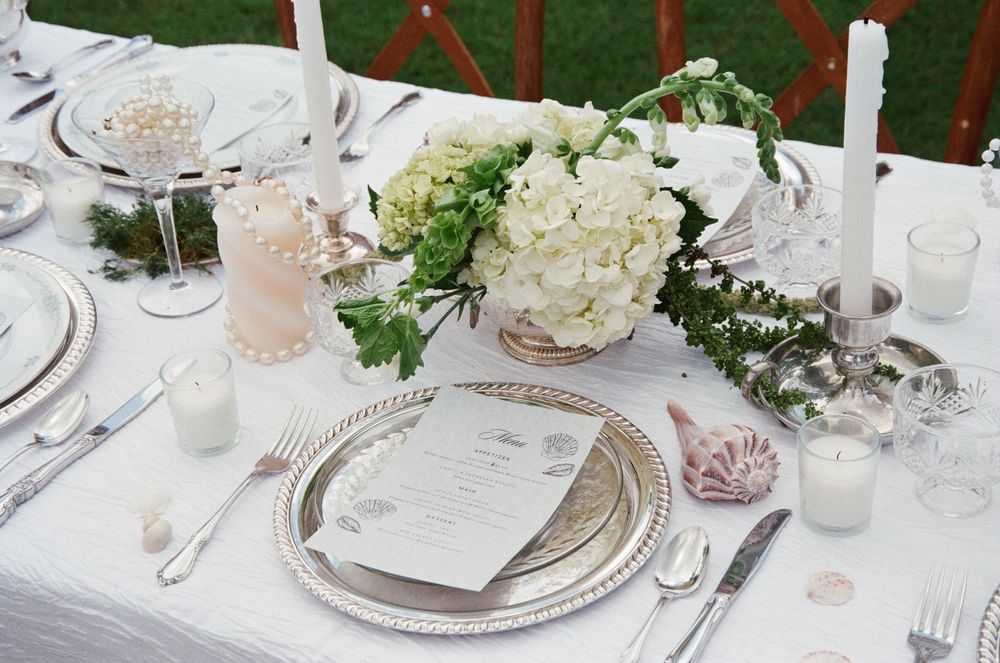 Elegant wedding reception table settings with white flowers, crystal glasses, and silver charger plates arranged outdoors.