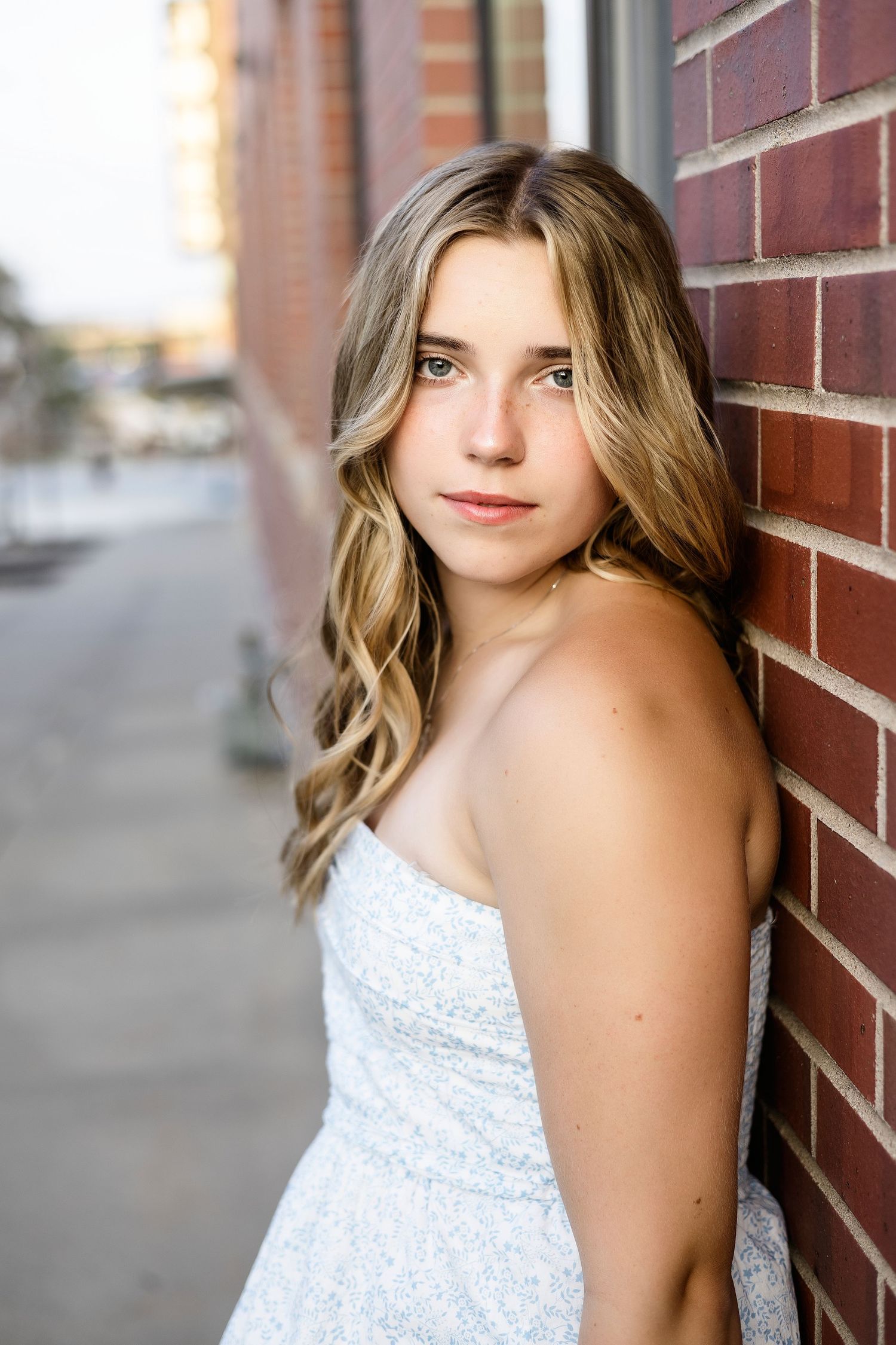 A woman in a white strapless dress poses against a brick wall on a city street at sunset.