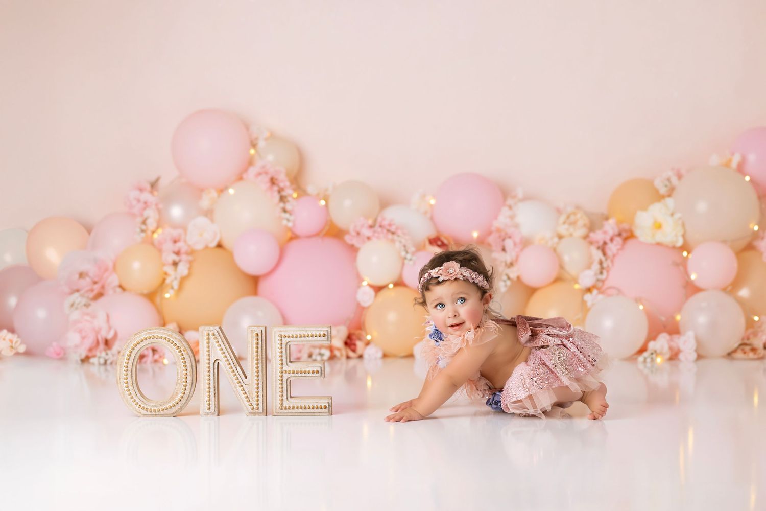 Baby sitting near letter ONE sign with pink and peach balloon arch decoration in studio setting.