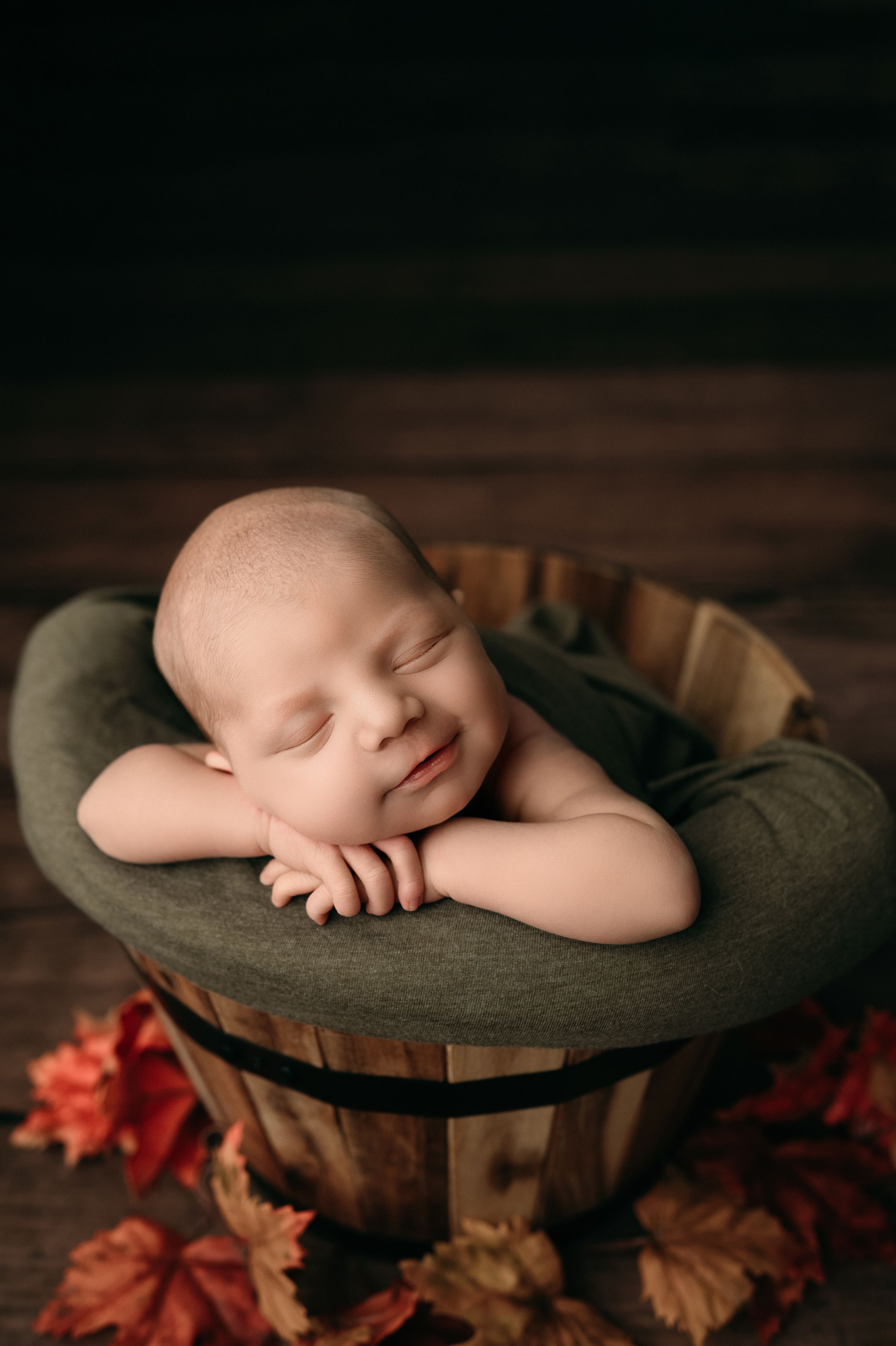 Sleeping baby nestled in dark green cushioned bowl with autumn leaves scattered around on wooden surface.