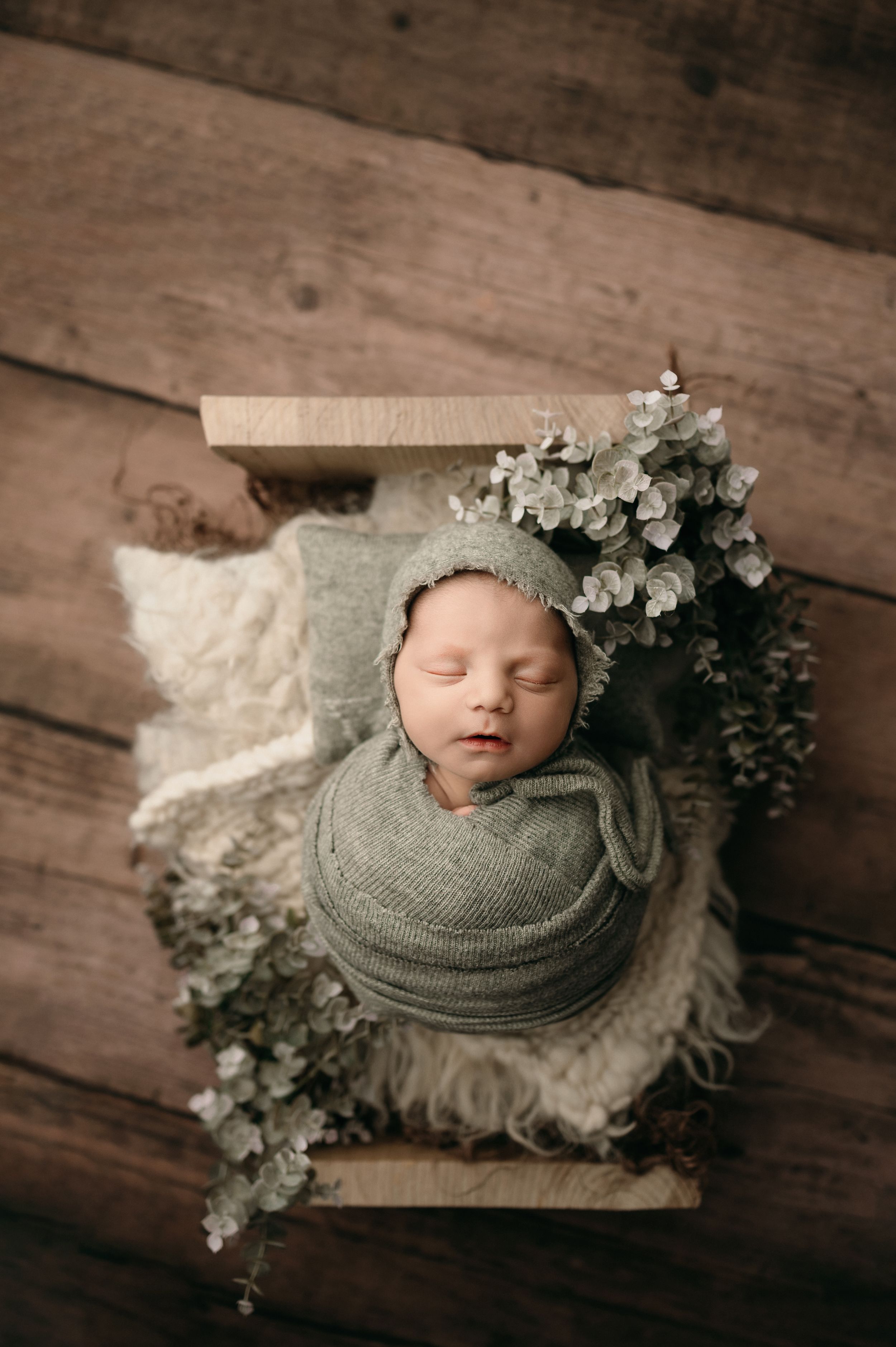 A newborn wrapped in sage green swaddle rests peacefully on fluffy fabric with eucalyptus sprigs in a wooden box setting.