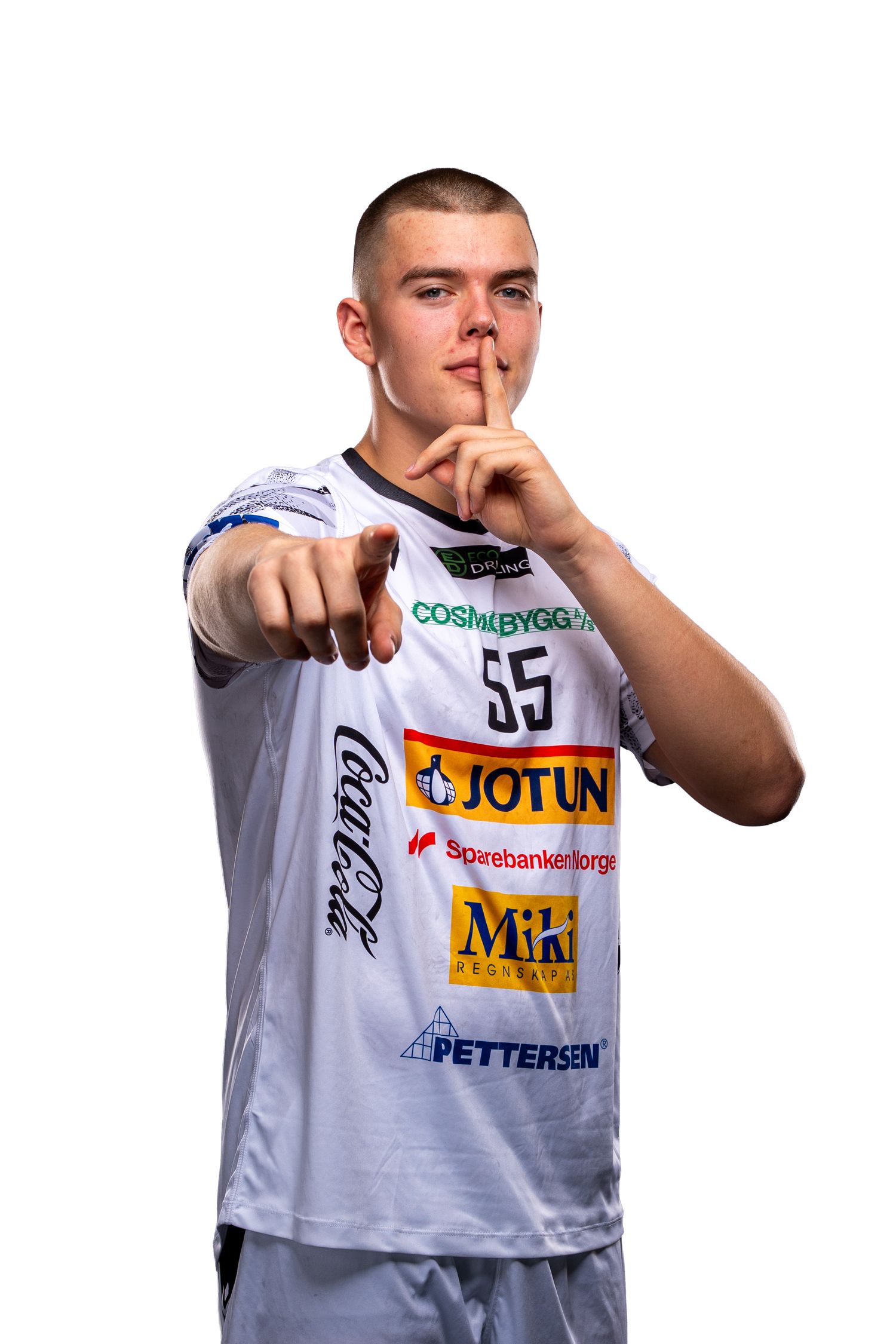 Athlete in white jersey with sponsor logos pointing forward while making shushing gesture with finger to lips.