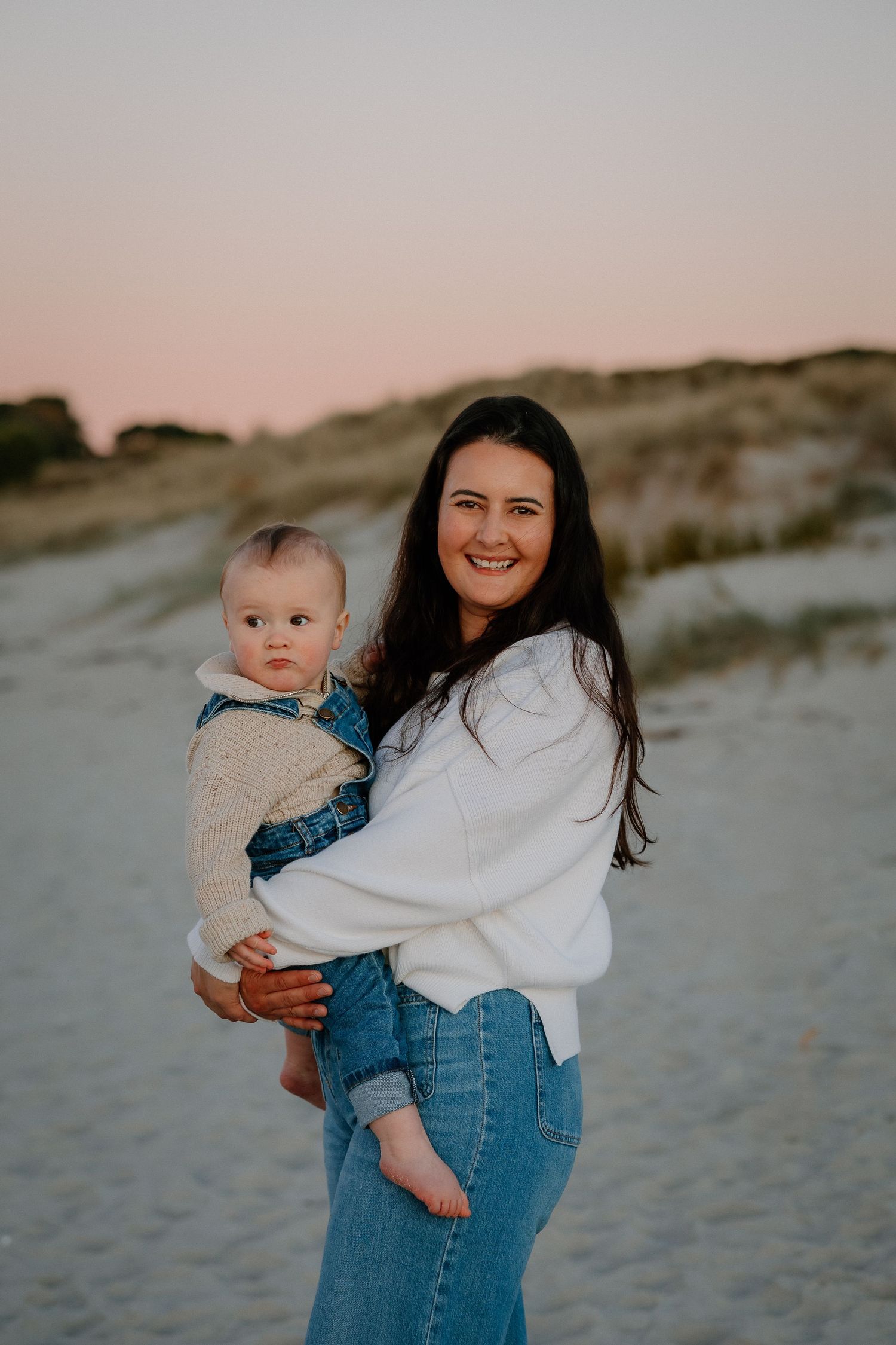 Parent and baby in casual denim and white outfits pose together on a sandy beach at sunset.