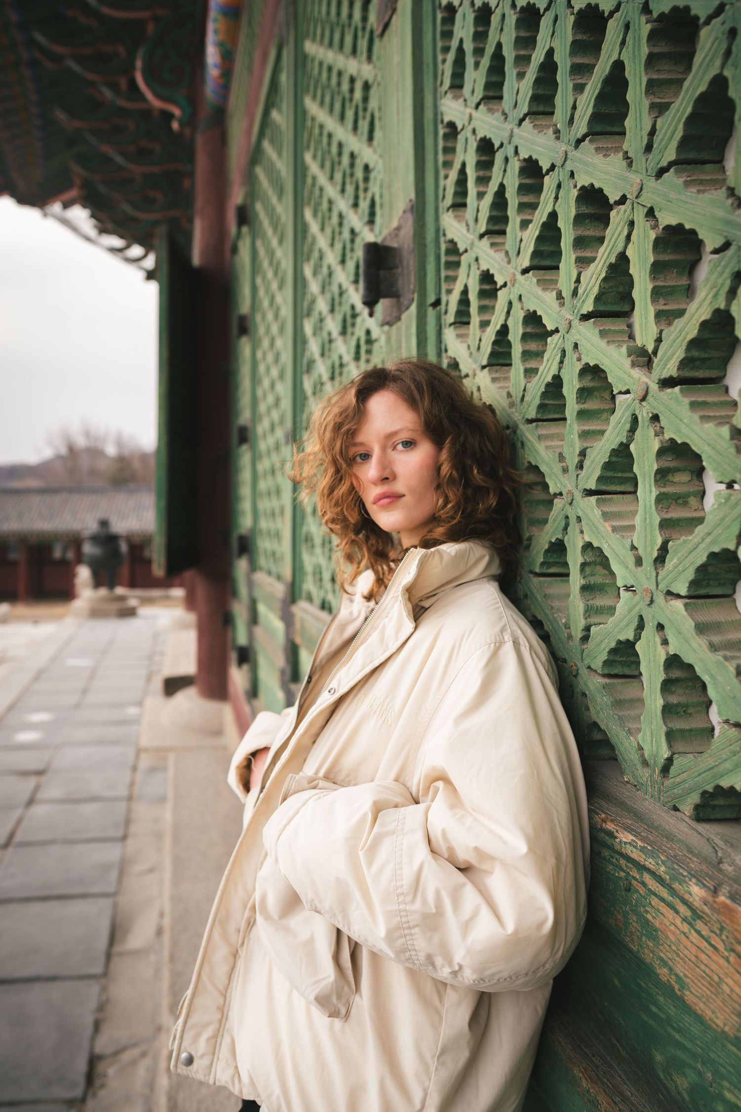 Person in a cream puffer jacket poses against ornate green architectural details.