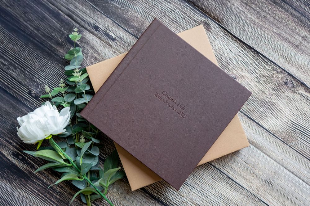 Brown leather wedding photo albums and envelopes arranged with green foliage on rustic wooden plank background.