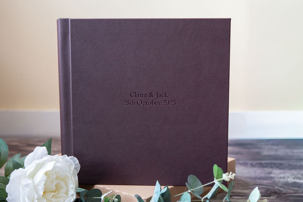Elegant brown leather wedding album displayed with white roses and rustic decor elements on wooden surface.