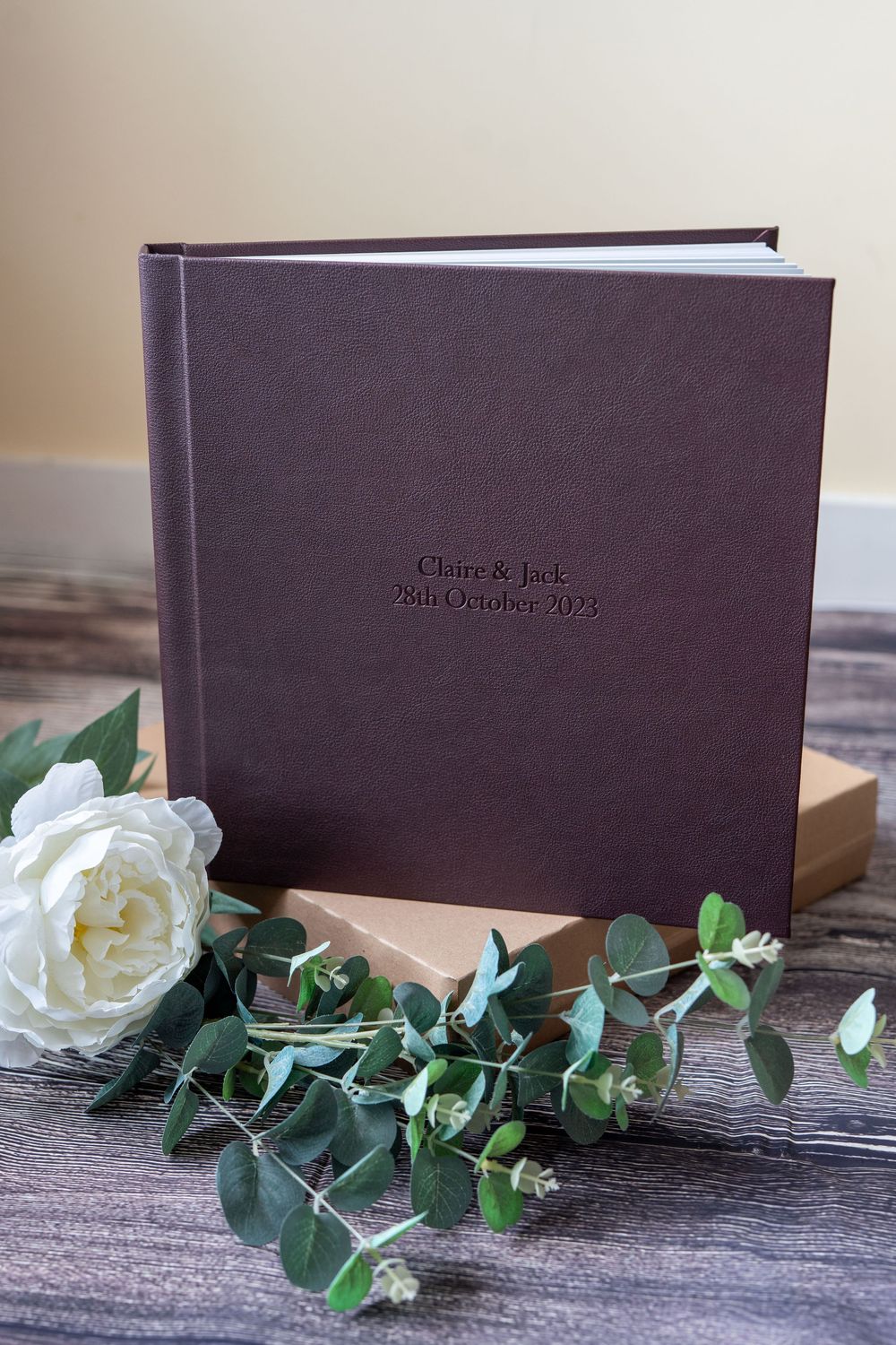A brown leather photo album or book displayed with white roses and eucalyptus leaves on a rustic wooden surface.