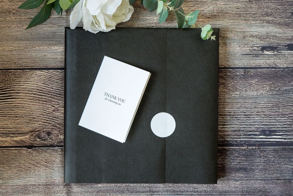 White rose petals and a black leather portfolio lay arranged on rustic wooden planks with small paper cards.