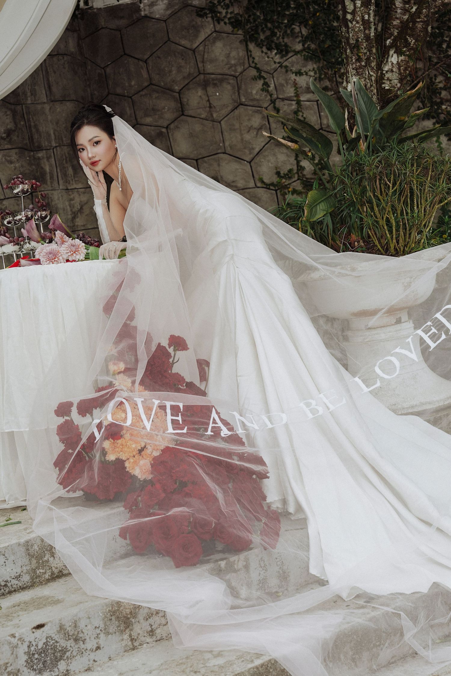 Beautiful Asian bride in flowing white wedding gown and long veil posed on stone steps with red roses.
