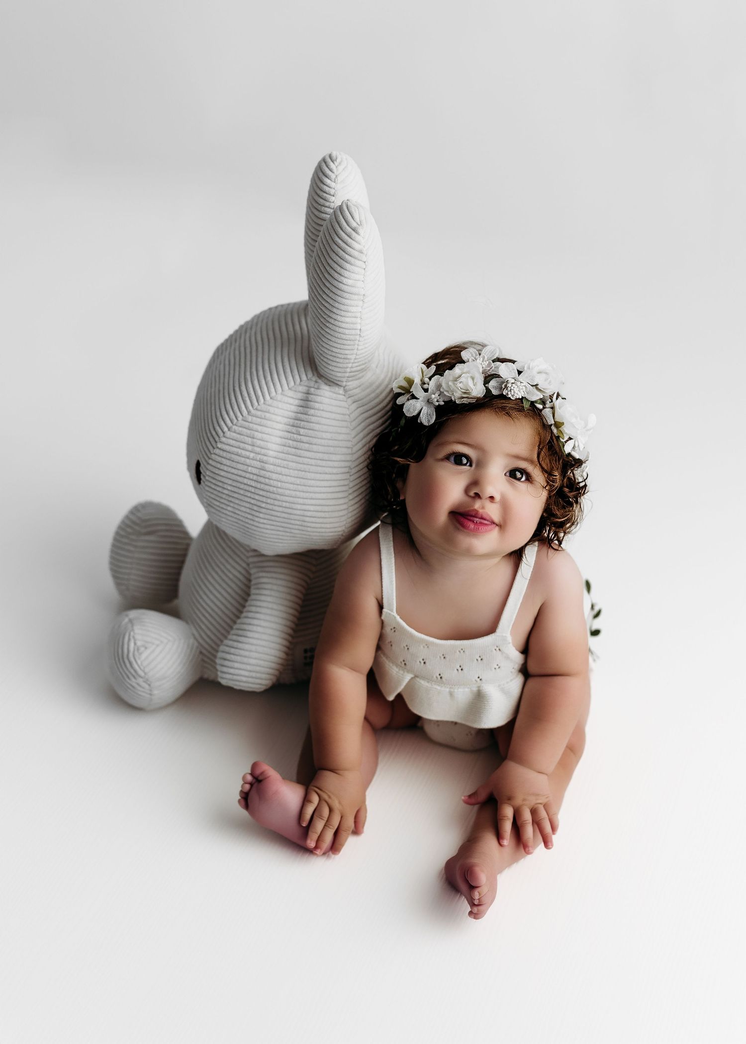 Baby wearing floral crown poses next to large white decorative bunny sculpture against white background.