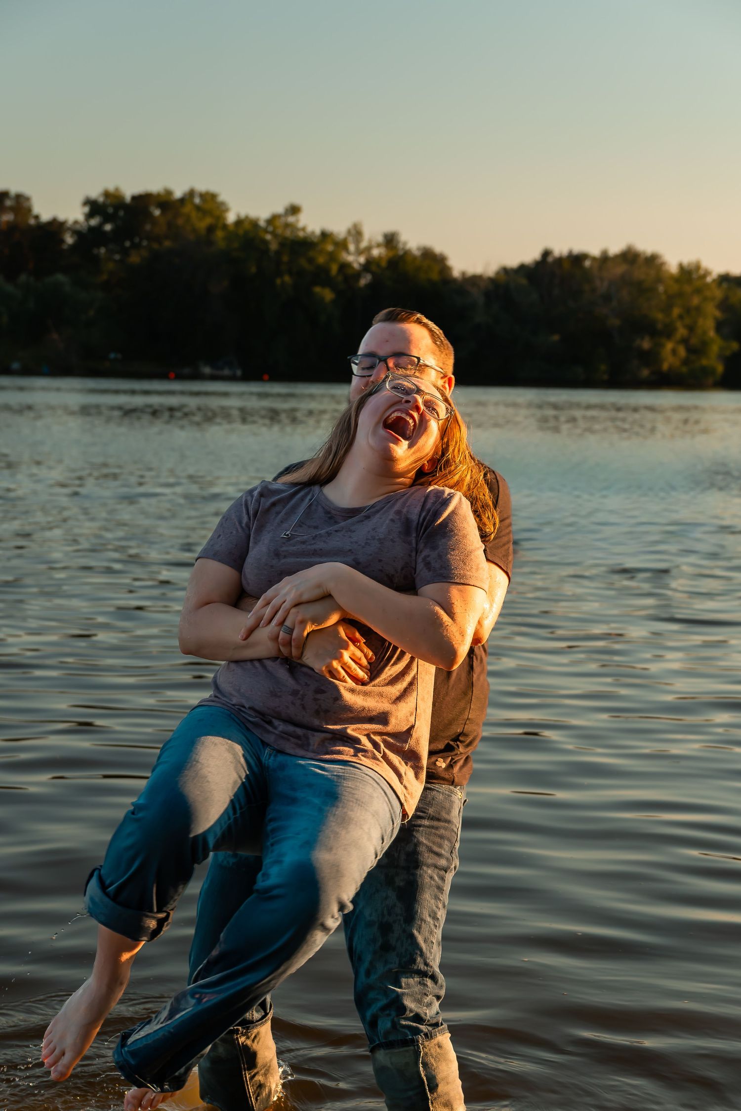Couple laughing joyfully at sunset by a lake, woman lifted in a playful embrace near the water.