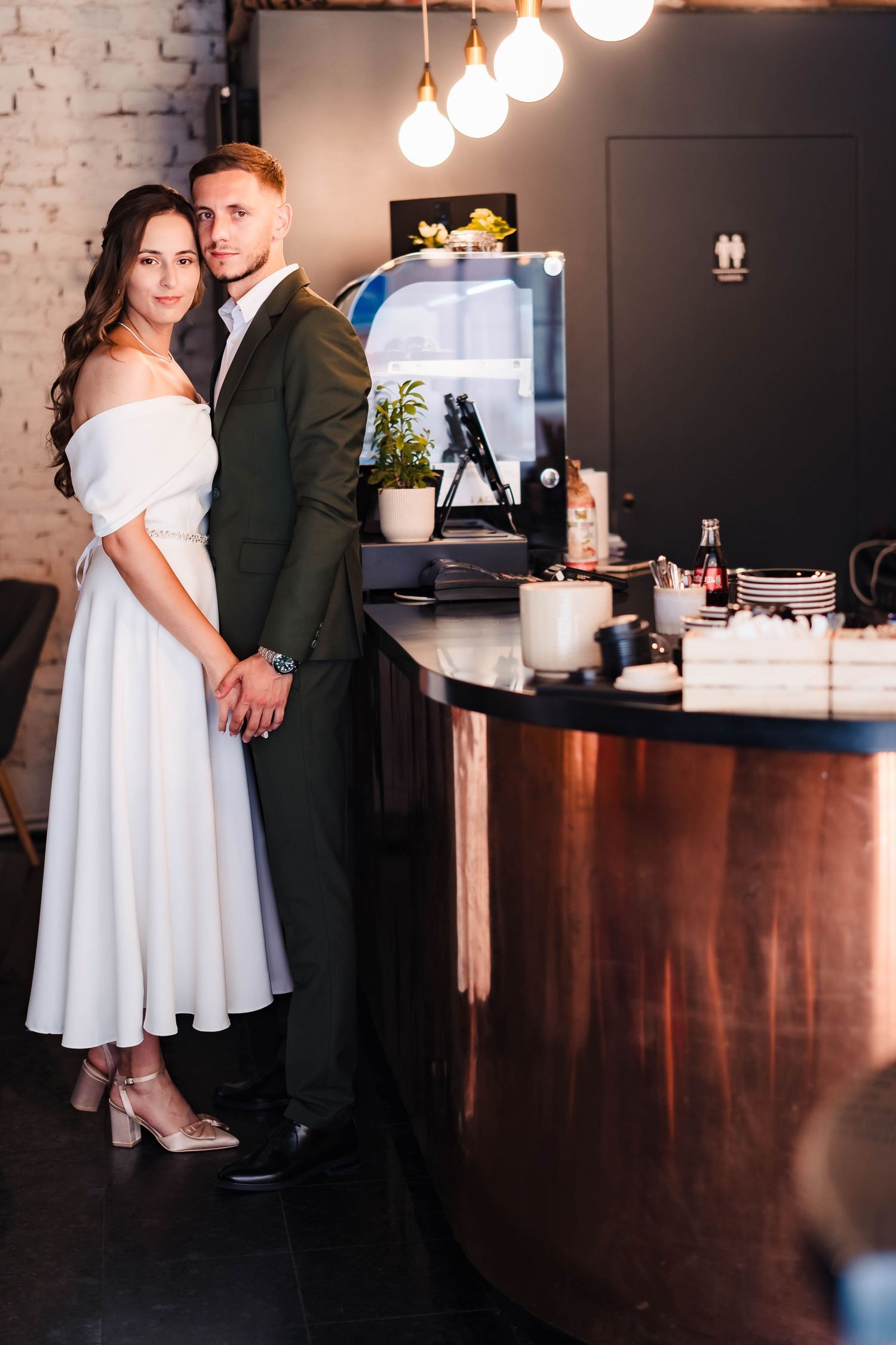 Couple shares intimate moment at bar counter with white dress and dark suit in moody lighting.