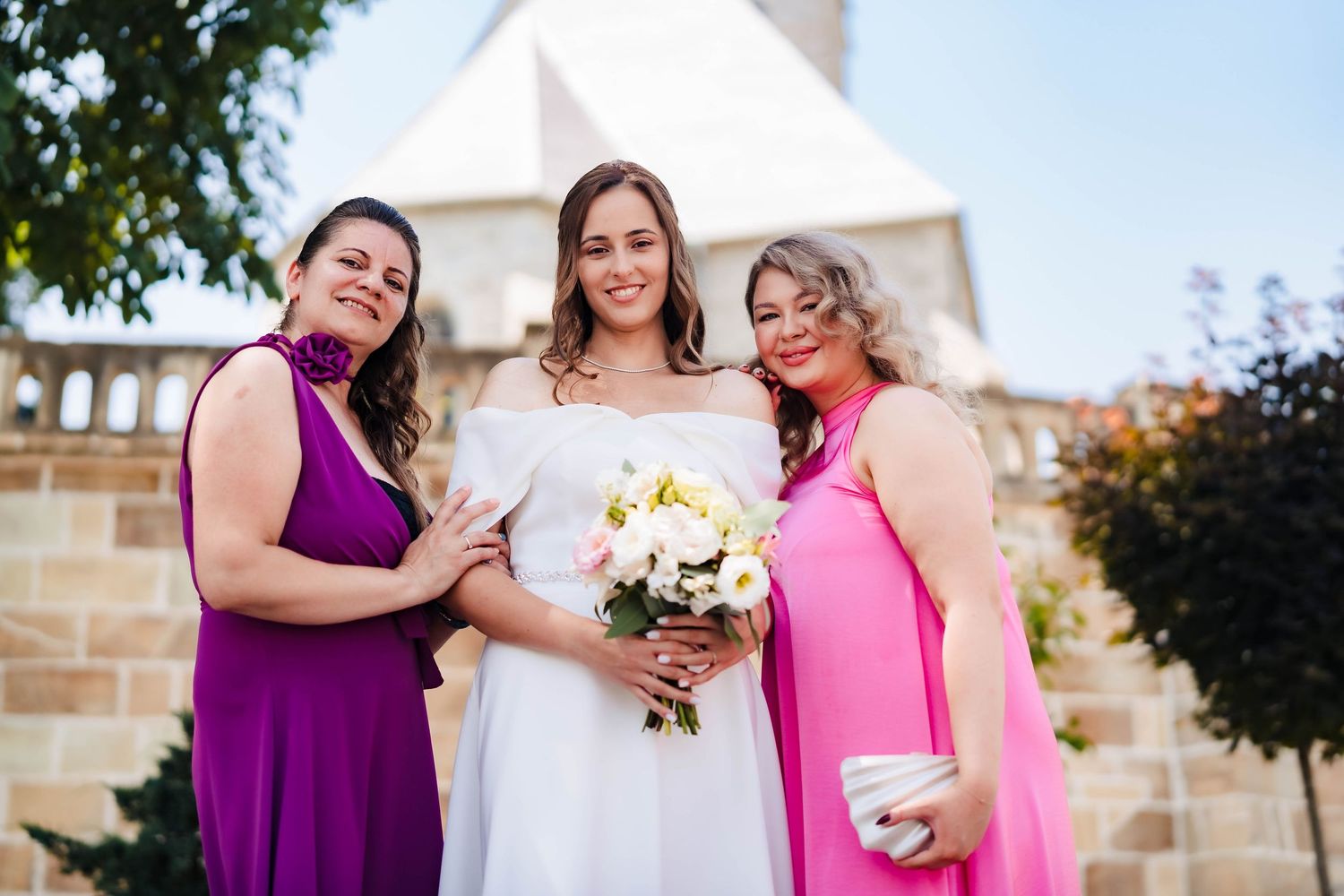 Wedding party members in purple and pink dresses pose outdoors with floral bouquets.