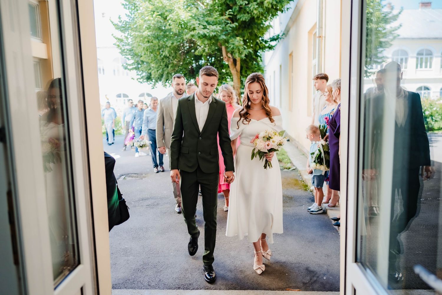 Wedding party walks down a tree-lined path outside a stone building.