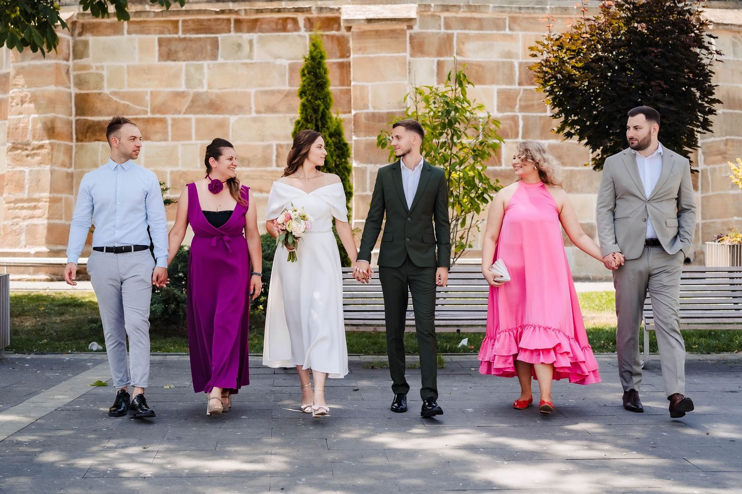 Wedding group photo session with coordinated outfits in pink and purple against stone wall.