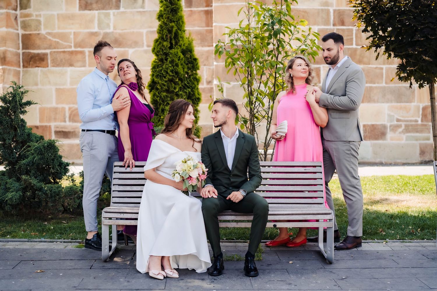 Wedding party relaxes on benches against a stone wall backdrop.