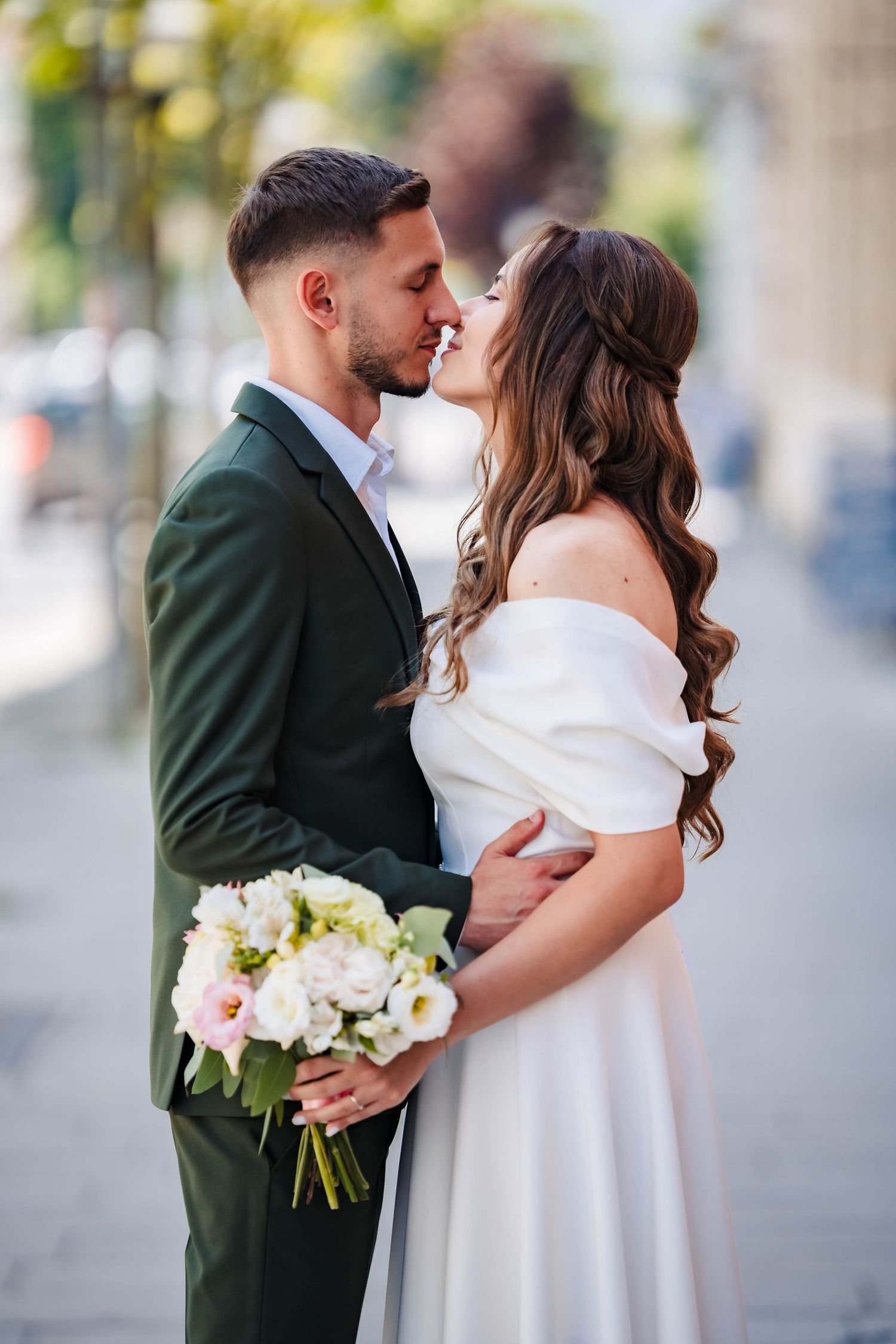 A couple in wedding attire share an intimate moment on a city street with white bouquet.