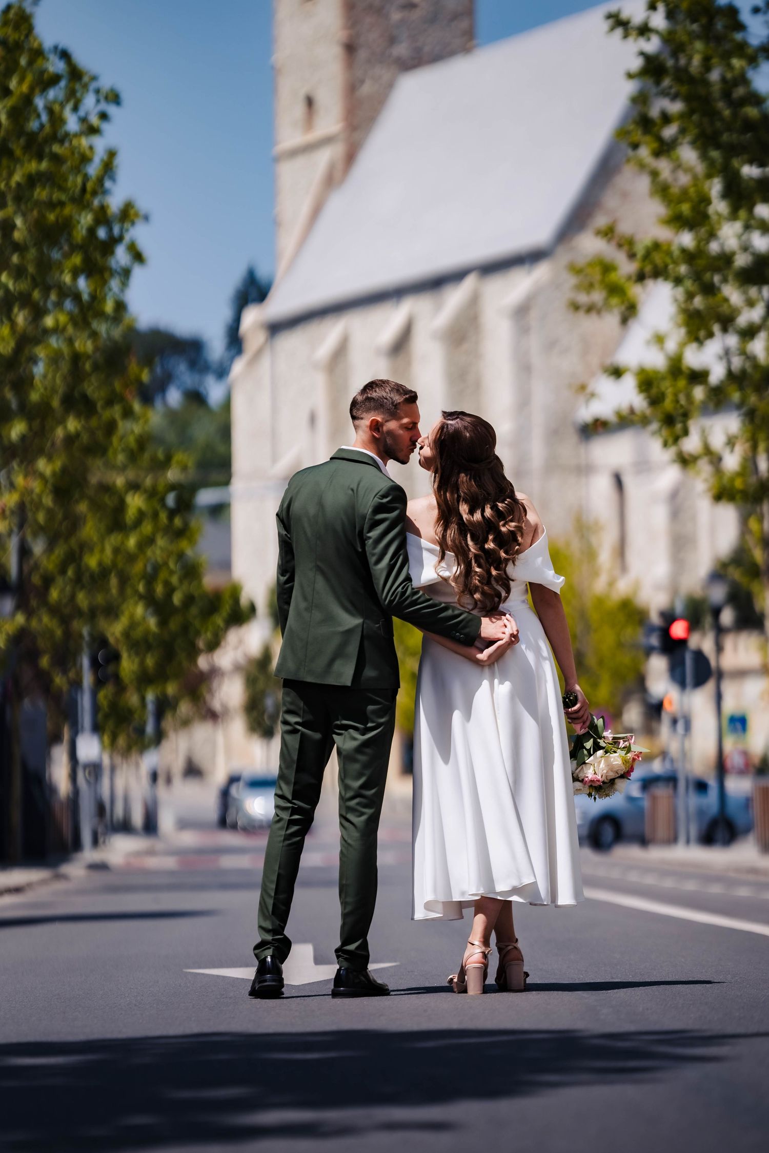 A romantic wedding couple poses on a street with a church building in the background.