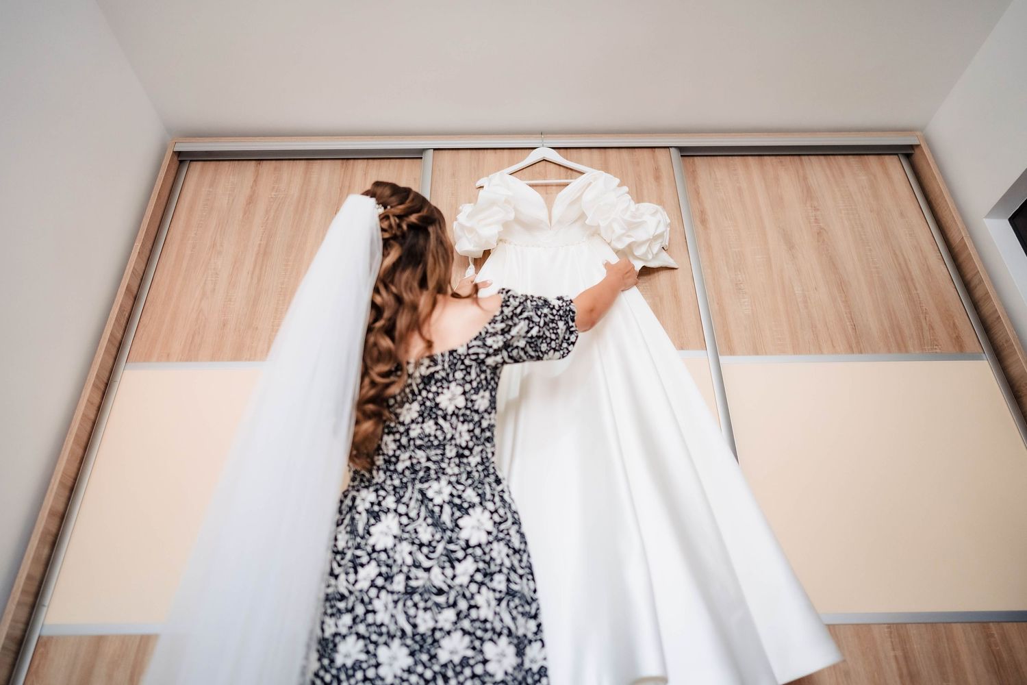 A white wedding dress hangs elegantly on a wooden hanger against a light wall.