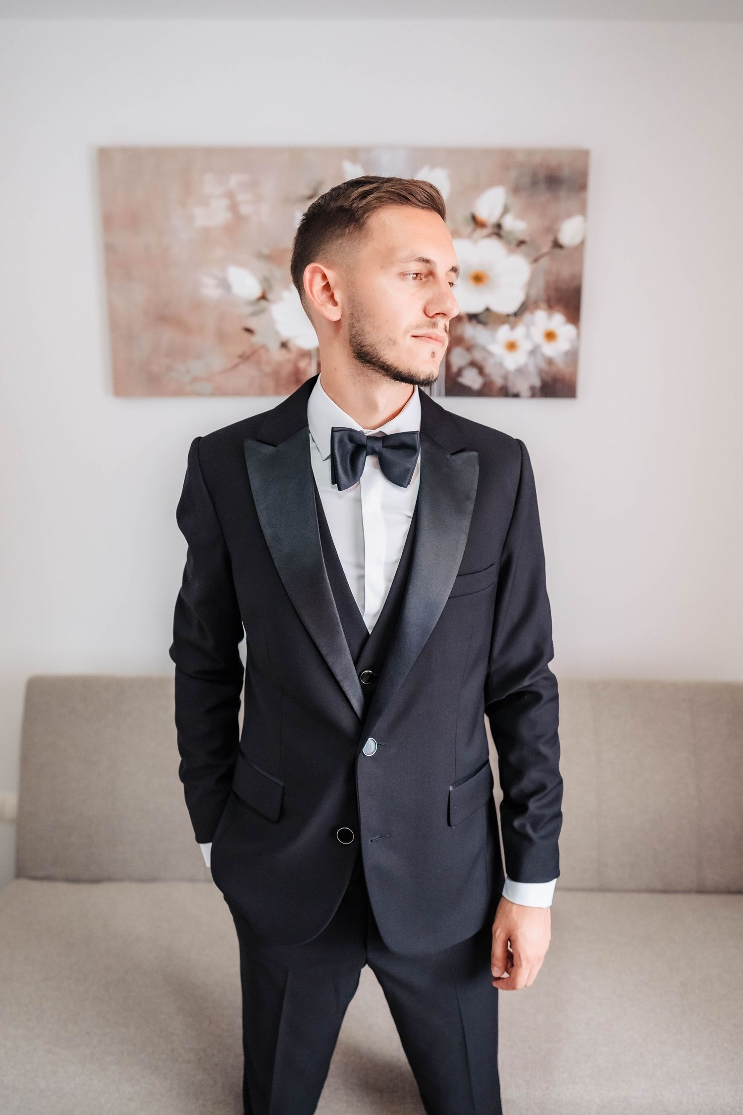 Groom in classic black tuxedo and bow tie poses in modern interior setting.