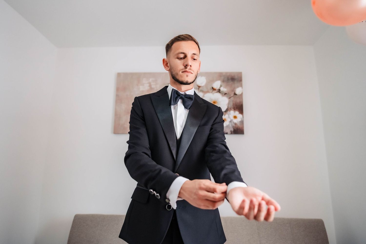 Groom in black tuxedo smiles while adjusting his bowtie before the ceremony.