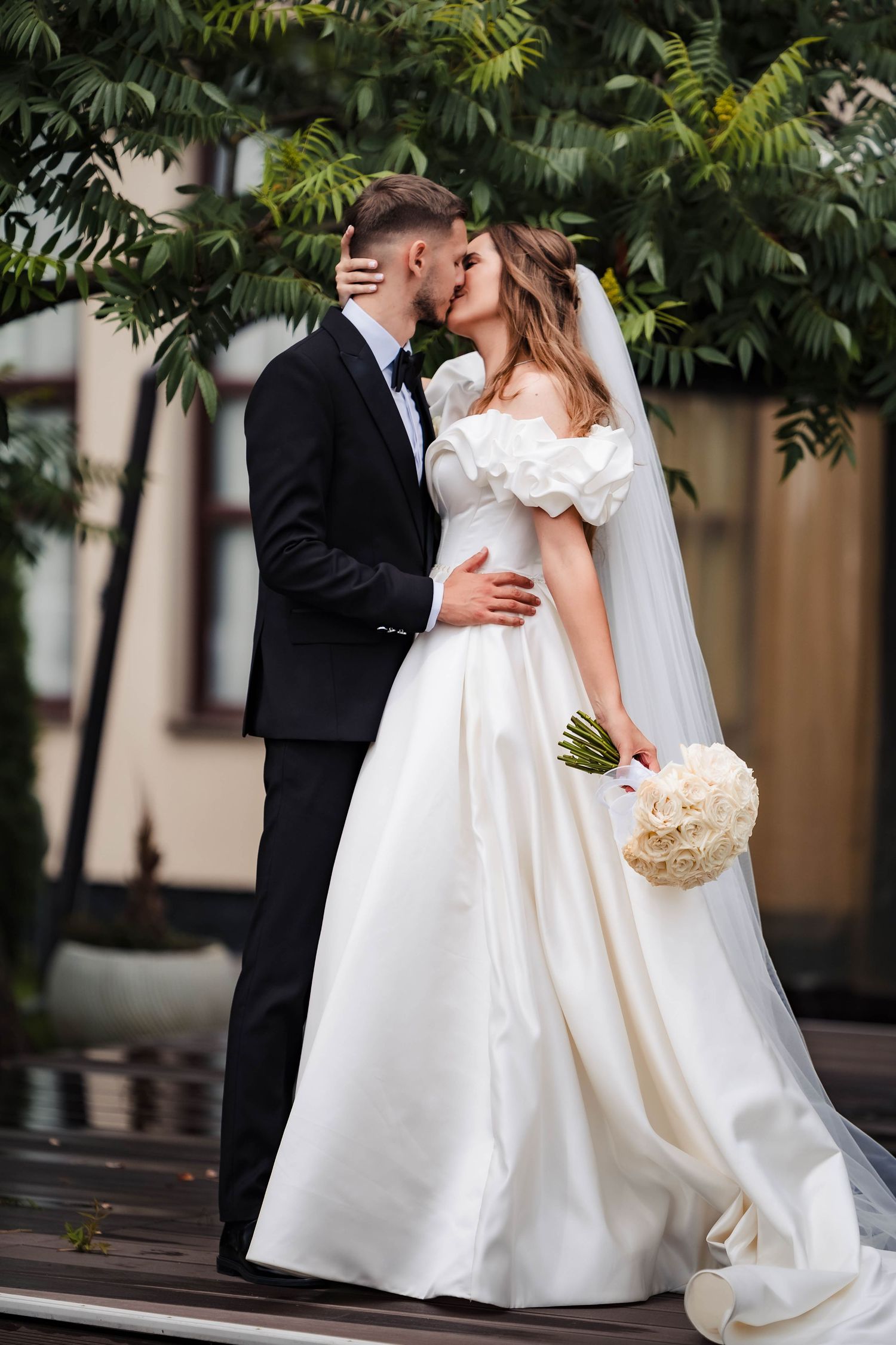Romantic wedding couple poses outdoors with lush greenery and white floral bouquet.