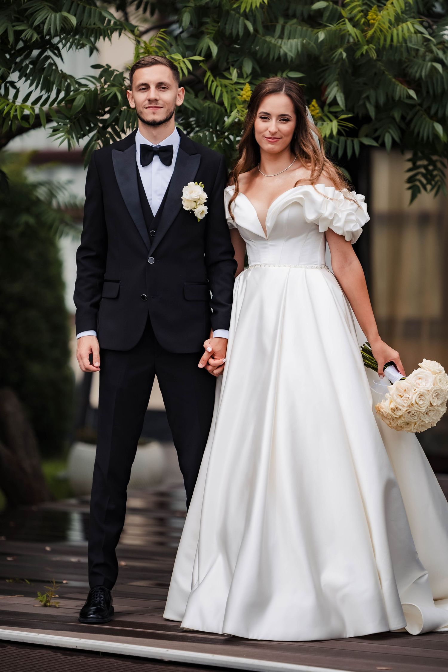 Newlyweds walking together outdoors, bride in flowing white gown and groom in classic tuxedo.