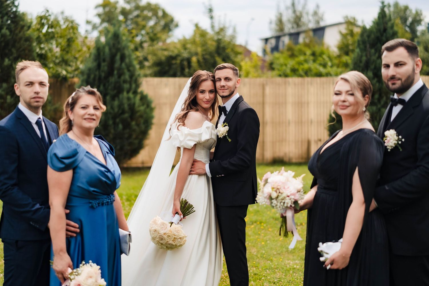 Wedding party poses outdoors in formal attire with bridesmaids in blue dresses and groomsmen in dark suits.