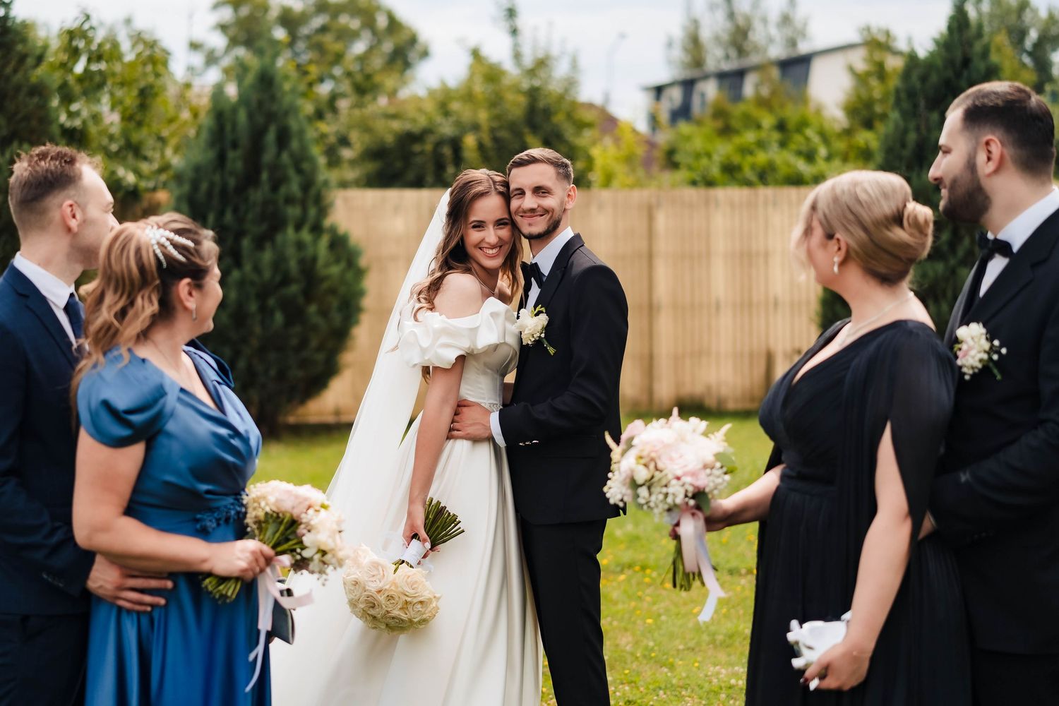 Wedding party gathered outdoors for photos on a sunny day with natural greenery.