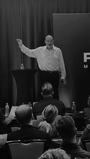 A speaker gesturing to an audience in a dark room during a presentation.