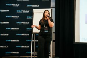Speaker presents on stage at business event with branded Eventpreneur backdrop panels visible.