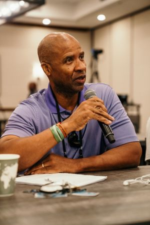 Man in blue polo shirt sits at conference table during business meeting while speaking into microphone.