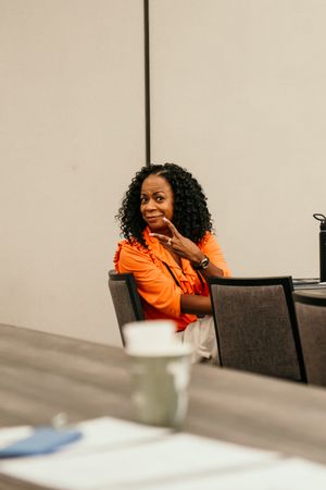 Person in bright orange hoodie sits at conference table appearing to be laughing or reacting.