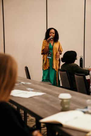 A person in a brown jacket and green dress gestures animatedly while speaking at a business meeting.
