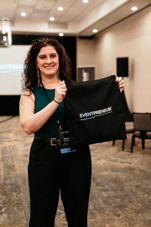 Event staff member displays black uniform shirt in conference room setting with carpeted floor and recessed lighting.