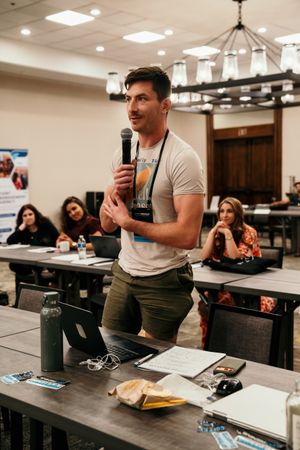 Speaker stands on table in classroom setting during presentation with audience seated at tables in background.