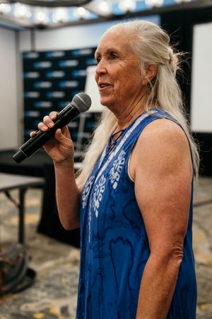 An elderly person in a blue patterned dress speaks into a microphone at an indoor event.
