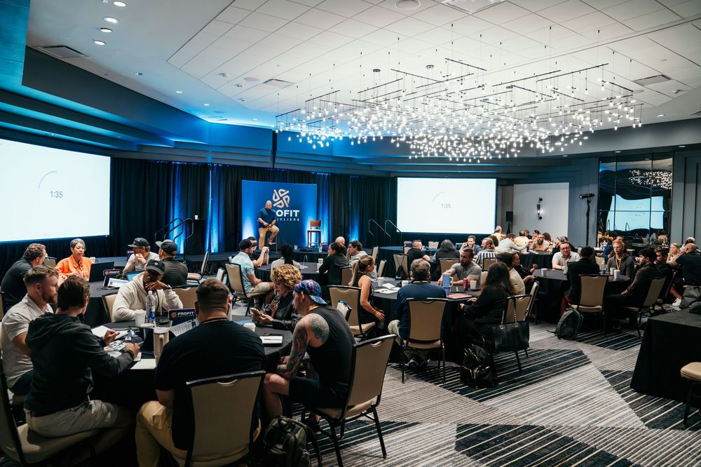 A large conference room filled with attendees during a business presentation with blue lighting and projection screens.