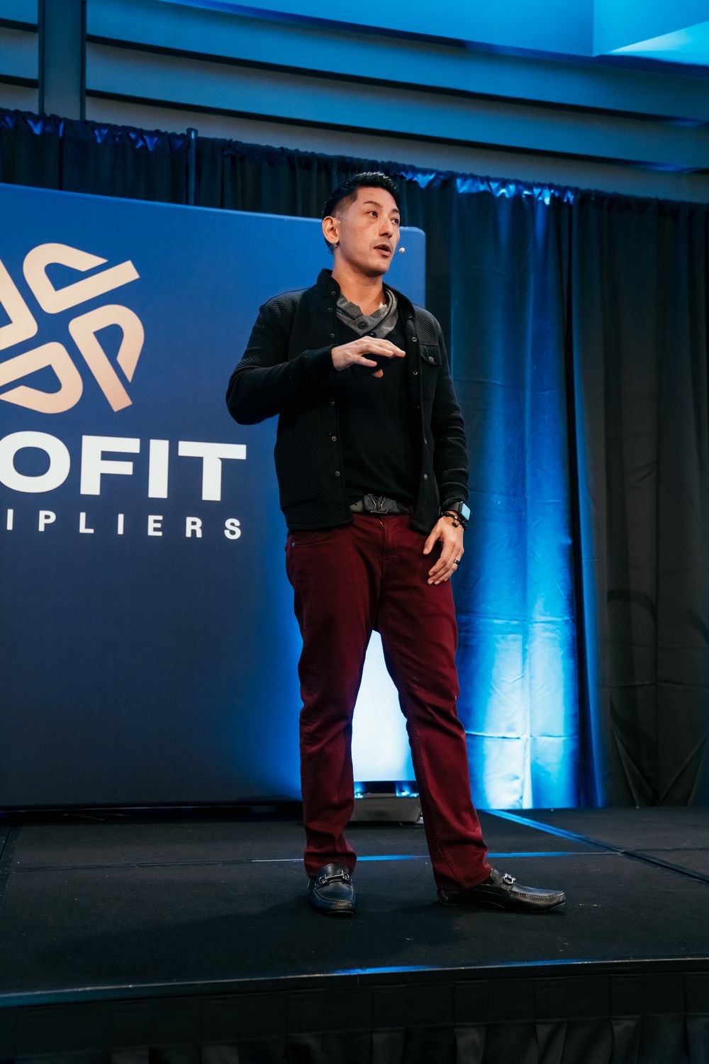 A speaker in black jacket and burgundy pants gestures dramatically on a blue-lit stage with corporate branding.