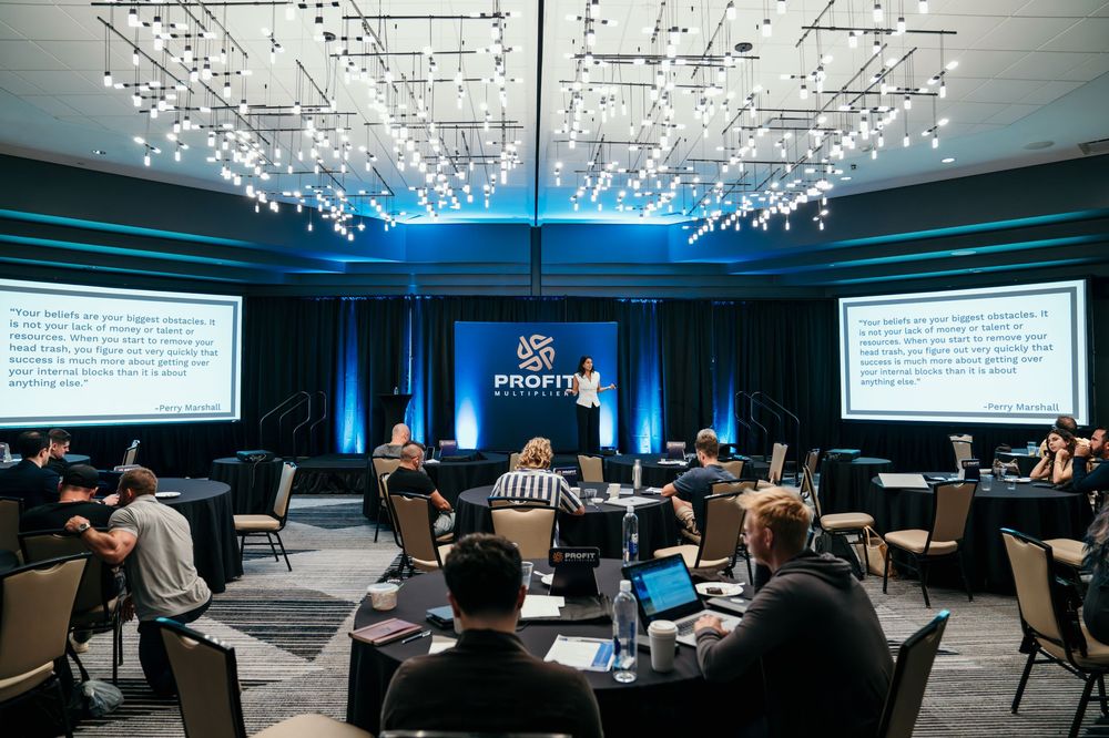 Conference attendees seated at round tables in a modern venue with crystal chandeliers and large presentation screens.