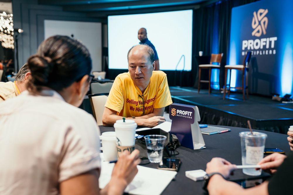 People gathered around a table having a discussion at an event with Profit Fitness branding in the background.