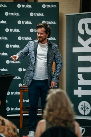 A speaker in a patterned blazer gestures while presenting at a Truka branded corporate event.
