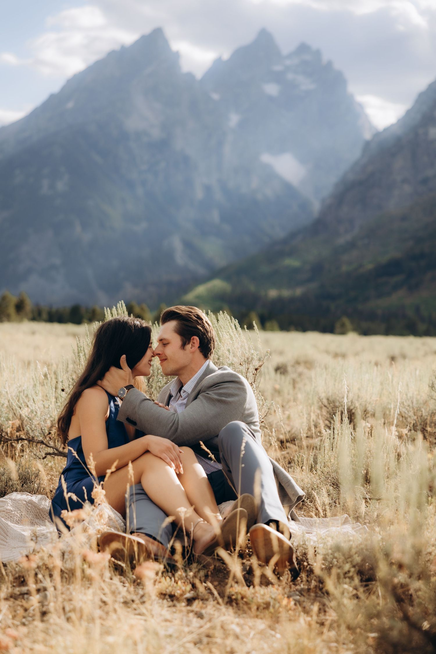 A loving embrace shared in a golden field with majestic mountains in the background at sunset.