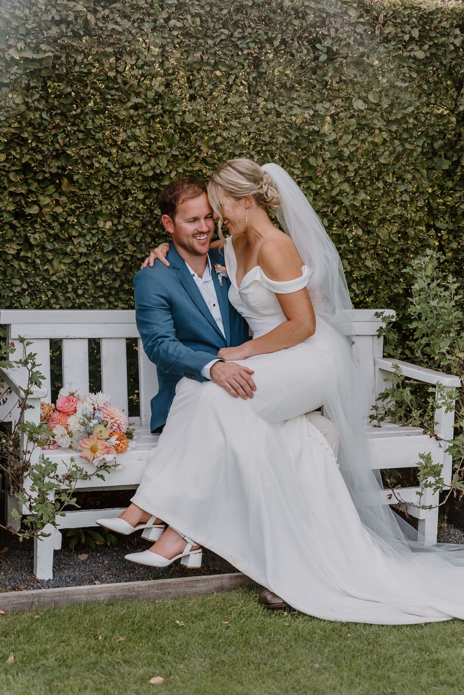 A romantic wedding couple sits together on a white bench surrounded by lush greenery. christchurch wedding photography