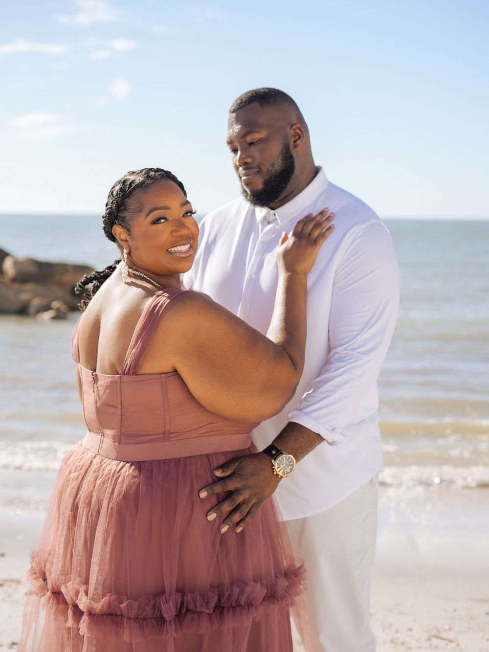 A couple embraces on a sunny beach as they pose for romantic maternity photos in a flowing mauve dress.