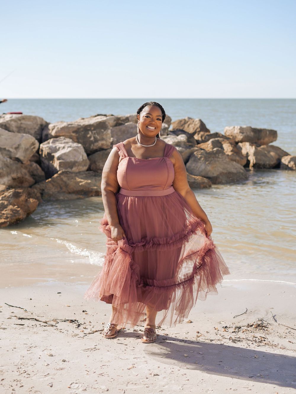 A woman in a flowing dusty pink dress poses joyfully on a rocky beach shoreline.