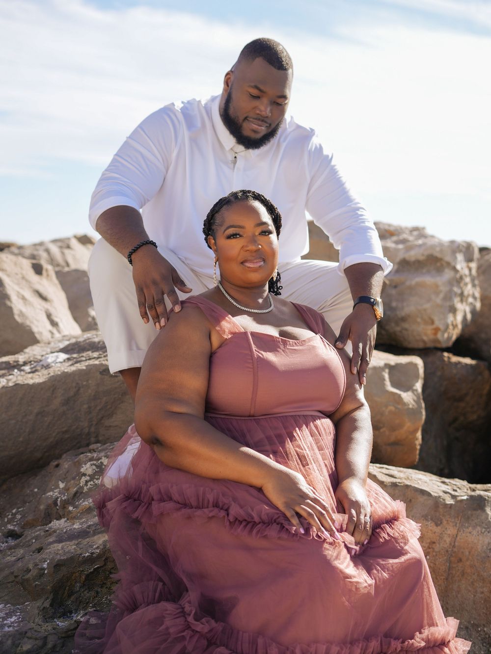 A couple poses together on rocky terrain during a romantic outdoor photoshoot, with the person in a flowing mauve dress.