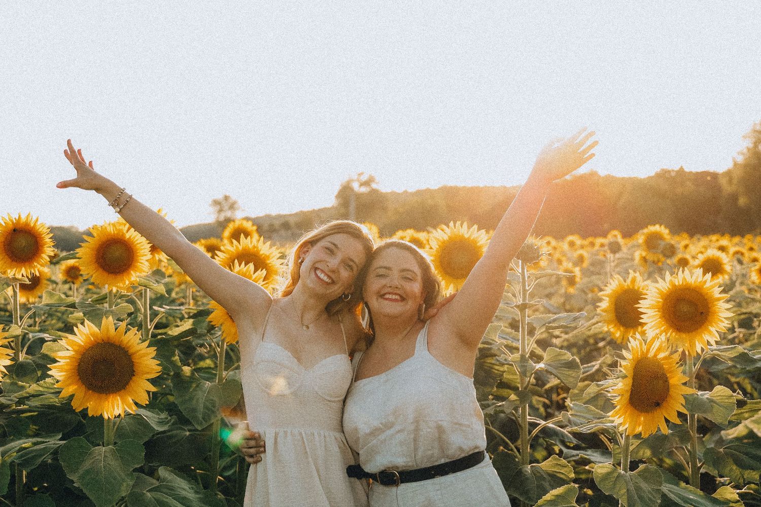 Two joyful women in white dresses raising arms in sunny sunflower field.