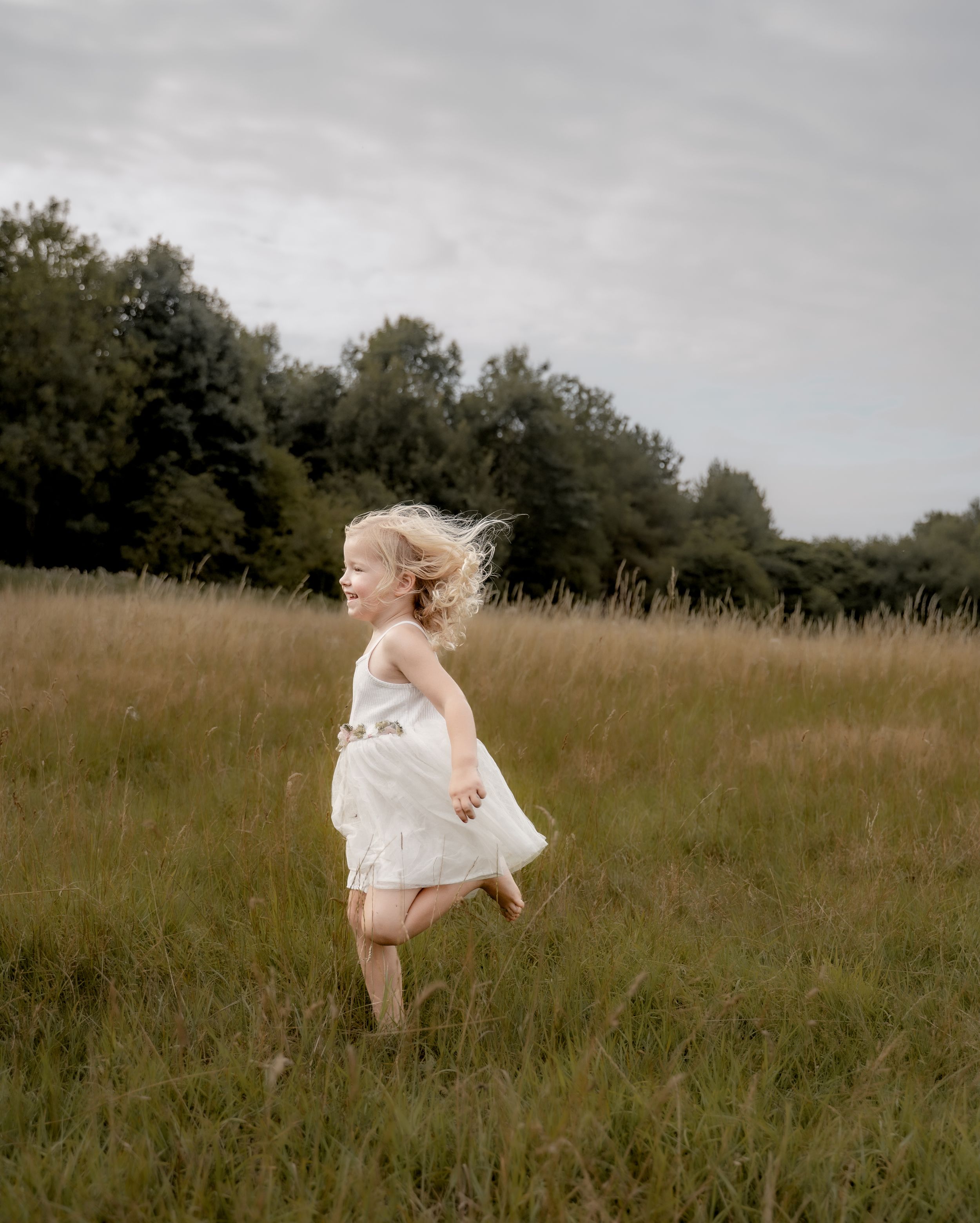 A person in a flowing white dress runs through a sunlit meadow of tall grass on a cloudy day.